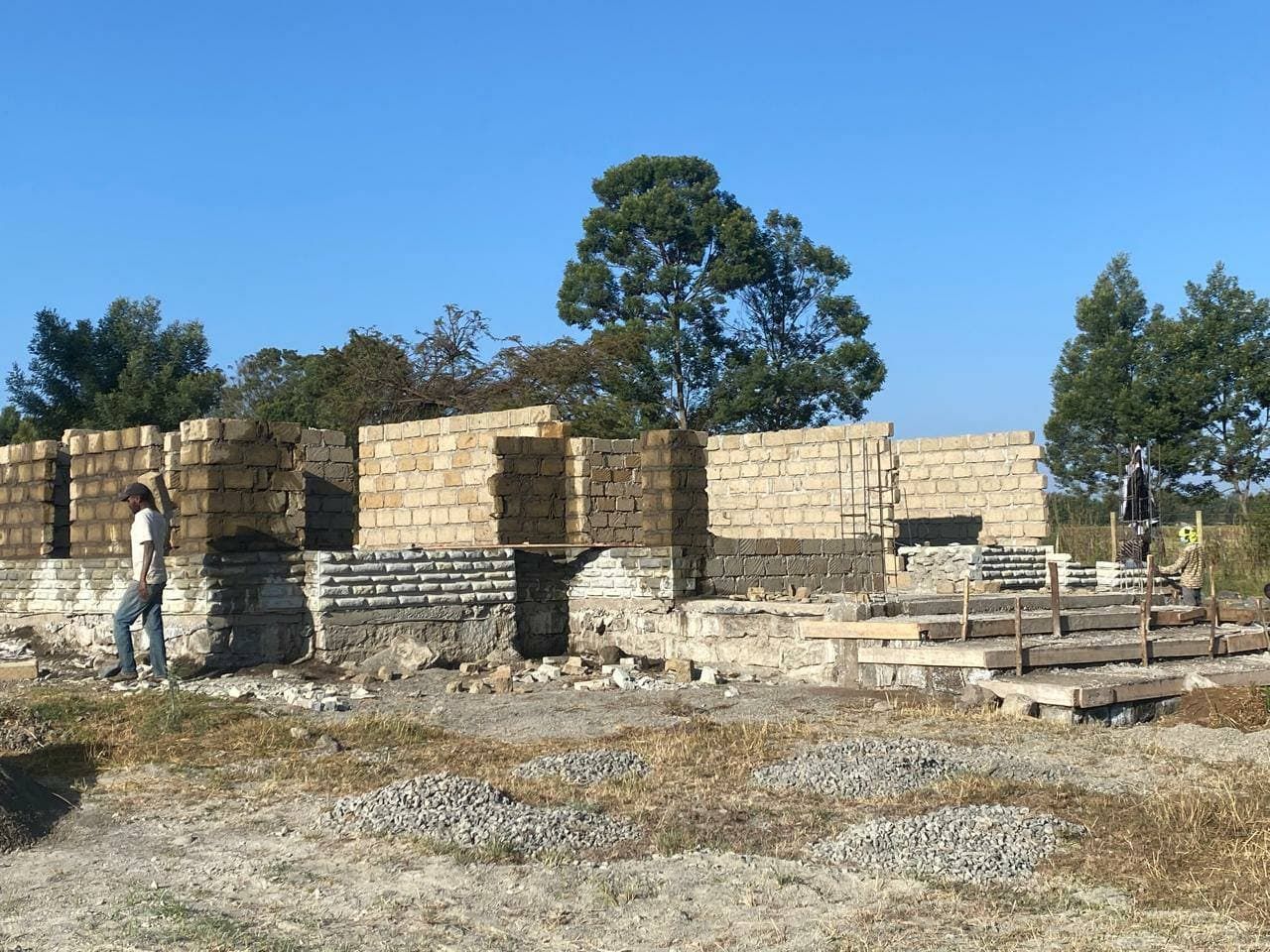 Quarry-block masonry walls under construction on a stone foundation, with timber formwork positioned for a concrete ring beam
