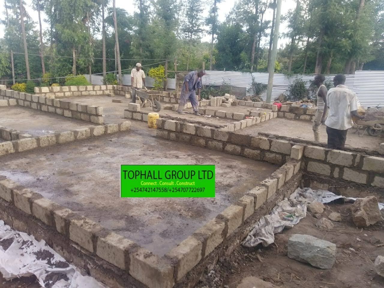 Construction site showing natural stone strip foundations built to floor level, with workers moving materials by wheelbarrow